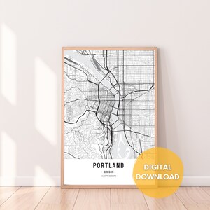 Op de afbeelding: Een zwart-witte kaartprint van Portland, Oregon met de naam van de stad en de coördinaten onderaan. De kaart is ingelijst in een licht houten lijst en wordt weergegeven tegen een witte muur. De tekst "DIGITAAL DOWNLOADEN" staat in een gele cirkel.