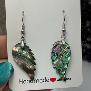 Puede incluir: Pendientes en forma de alas, con un acabado brillante e iridiscente. Los pendientes muestran tonos de verde, morado y dorado. Cuelgan de ganchos plateados. La tarjeta dice "Handmade with love."