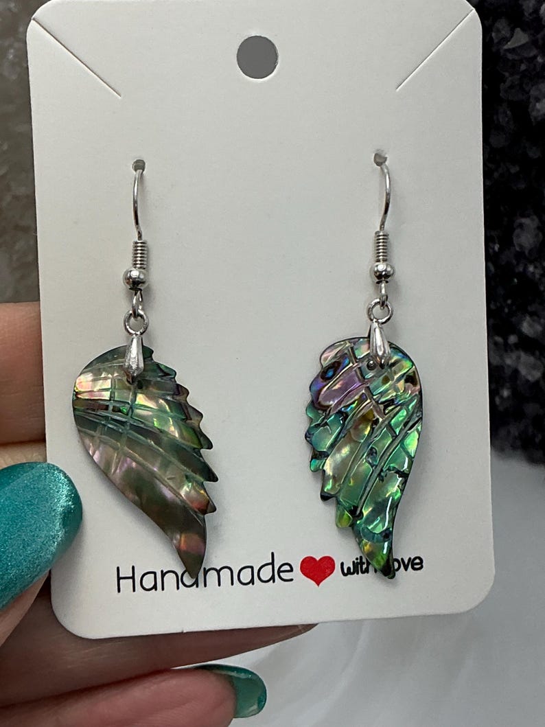Puede incluir: Pendientes en forma de alas, con un acabado brillante e iridiscente. Los pendientes muestran tonos de verde, morado y dorado. Cuelgan de ganchos plateados. La tarjeta dice "Handmade with love."