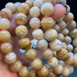 May include: A multi-strand bracelet composed of polished, spherical beads. The beads feature a banded, translucent appearance with swirling patterns in shades of cream, beige, tan, and soft orange, creating a natural, layered stone aesthetic.