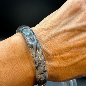 May include: A bracelet composed of smooth, rectangular, polished rutilated quartz beads. The beads are translucent with visible black needle-like inclusions throughout the stone, strung together to form a continuous circular band.