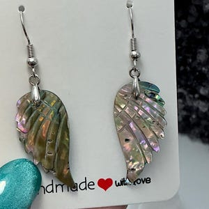 Puede incluir: Un par de pendientes iridiscentes en forma de ala con ganchos plateados. Los pendientes est&aacute;n hechos de un material similar a una concha con tonos verdes, rosas y morados. La tarjeta detr&aacute;s de los pendientes dice "handmade with love".