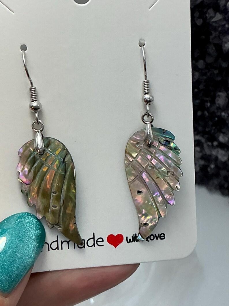 Puede incluir: Un par de pendientes iridiscentes en forma de ala con ganchos plateados. Los pendientes est&aacute;n hechos de un material similar a una concha con tonos verdes, rosas y morados. La tarjeta detr&aacute;s de los pendientes dice "handmade with love".
