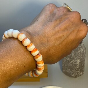 May include: A stretch bracelet composed of smooth, round beads featuring a white base color with vibrant orange marbled swirls throughout each bead. The beads are uniform in size and strung together to form a circular band that fits around the wrist.