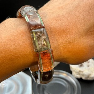 May include: A stretch bracelet composed of rectangular, polished beads in varying shades of translucent amber, deep brown, and mossy green with internal inclusions, worn around a wrist to showcase the smooth, rounded edges of each bead.
