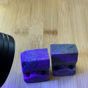Peut inclure: Deux petites pierres grises en forme de cube avec des visages sculptés. Les pierres ont des yeux noirs et des bouches souriantes. Les pierres sont sur une surface en bois brun clair et sont éclairées par une lumière violette.
