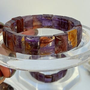 Puede incluir: Un brazalete rectangular de amatista con una mezcla de tonos morados, marrones y dorados. El brazalete está hecho de piedras pulidas y facetadas. El brazalete se exhibe sobre un soporte circular transparente.