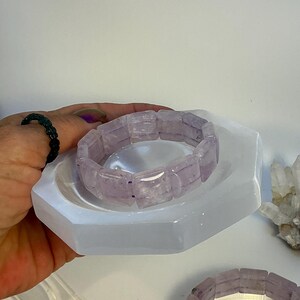Puede incluir: Una pulsera de amatista de color púrpura claro descansa sobre un plato de selenita blanco hexagonal. La pulsera está hecha de cuentas rectangulares de amatista. Una mano con un anillo de cuentas negro sostiene el plato. Cristales adicionales son visibles en el fondo.