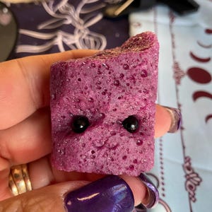 May include: A close-up of a purple, textured, square-shaped object with two black dots in the center. The object is being held by a hand.