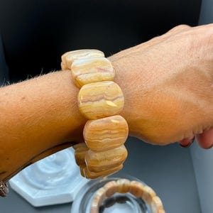 May include: A stretch bracelet composed of smooth, rectangular, polished beads featuring banded patterns in shades of cream, tan, and light orange. The beads have a glossy finish and are arranged in a single row, showing natural mineral variations.