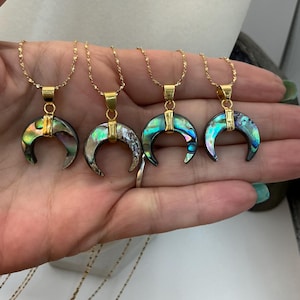 May include: Four gold necklaces, each with an iridescent crescent moon pendant. The pendants feature green, blue, and purple hues. The necklaces are presented against a light backdrop.