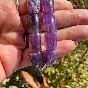 May include: Two amethyst bracelets with rectangular beads. The bracelets are made of natural amethyst stones and have a purple hue.