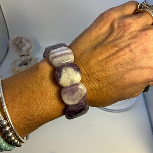 Puede incluir: Un brazalete con grandes piedras de amatista pulidas en tonos morados y blancos. El brazalete está hecho de varias piedras redondeadas ensartadas. Las piedras son lisas y tienen un aspecto veteado natural. El brazalete se lleva en una muñeca con otras pulseras de plata.
