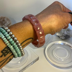 Puede incluir: Una colección de pulseras. Una pulsera de cuarzo rosa rojizo y facetado es el punto focal. Pulseras adicionales incluyen una pulsera de cuentas turquesa y una pulsera de cuentas plateadas. Las pulseras se muestran en una muñeca.