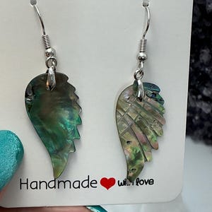 Puede incluir: Un par de pendientes iridiscentes en forma de ala con ganchos plateados. Los pendientes est&aacute;n hechos de un material brillante y multicolor, con tonos verdes, azules y rosas. La tarjeta dice "Handmade with love."