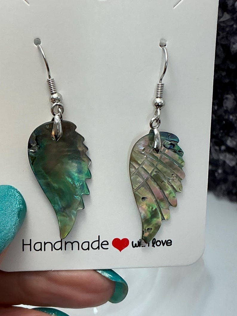 Puede incluir: Un par de pendientes iridiscentes en forma de ala con ganchos plateados. Los pendientes est&aacute;n hechos de un material brillante y multicolor, con tonos verdes, azules y rosas. La tarjeta dice "Handmade with love."