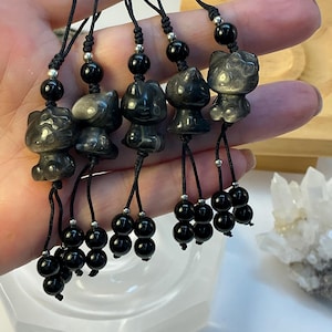 May include: Five black obsidian cat figurines on black cords, each with a cluster of black beads. The figurines are held in a hand, with a white bowl and crystal cluster in the background. The cat figurines have a glossy finish.