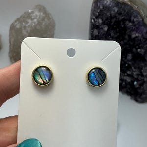 May include: Round stud earrings featuring iridescent abalone shells set in gold-coloured bezels. The earrings are presented on a white earring card. The card has the text "handmade with love".