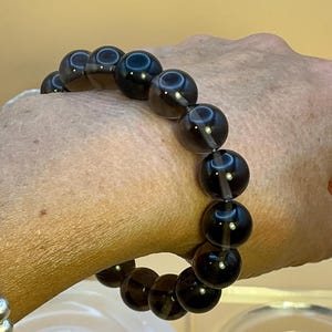 May include: A stretch bracelet composed of round, translucent dark brown beads. The beads have a smooth, polished surface that reflects light, creating small circular highlights across the dark, smoky-toned spherical stones arranged in a single row.