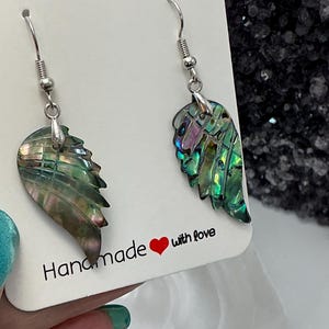Puede incluir: Un par de pendientes hechos a mano con forma de ala con conchas de abul&oacute;n iridiscentes. Los pendientes tienen ganchos plateados y se exhiben en una tarjeta blanca con el texto "Handmade with love".