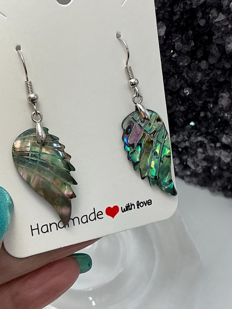 Puede incluir: Un par de pendientes hechos a mano con forma de ala con conchas de abul&oacute;n iridiscentes. Los pendientes tienen ganchos plateados y se exhiben en una tarjeta blanca con el texto "Handmade with love".