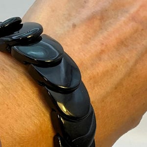 May include: A black bracelet composed of linked, glossy, oval-shaped segments. The bracelet is worn on a wrist, and the segments are interlocked to form a flexible band. The bracelet's design is simple and elegant, with a high-shine finish.