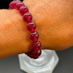 May include: A stretch bracelet composed of multiple smooth, round, translucent deep red beads. The beads are uniform in size and shape, featuring a polished, glossy finish that reflects light, strung together on an elastic cord to form a circular band.