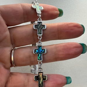 以下が含まれることがあります： 4つの十字架チャームが付いたシルバーブレスレット。各十字架には、緑、青、金などの色合いを示す、虹色のあわびの貝殻の象嵌が施されています。ブレスレットは小さなシルバーのリンクで接続されています。