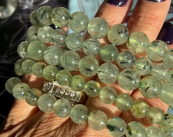 May include: A stack of green gemstone bracelets with black inclusions. The bracelets are made of round beads and are stacked on a person's wrist.