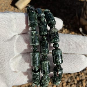 May include: Three strands of dark green gemstone beads, each strand is made of cylindrical beads with a speckled pattern. The beads are strung on a black cord.