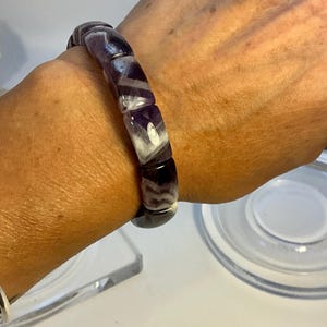 Puede incluir: Un brazalete con piedras rectangulares pulidas en tonos morados y blancos. El brazalete se lleva en la muñeca, mostrando las variaciones naturales de color y la textura suave de las piedras. El brazalete es una pieza de joyería.