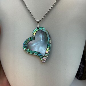 May include: A heart-shaped pendant necklace with a silver chain. The pendant features a pearlescent center framed by iridescent abalone shell. The abalone shell displays shades of green, blue, and purple. The necklace is displayed on a mannequin.