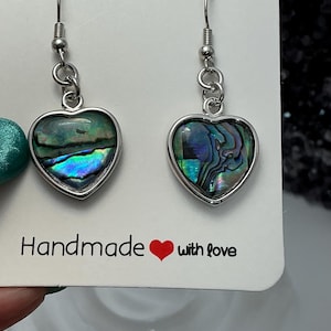 May include: Heart-shaped earrings with iridescent abalone shells set in silver-toned metal. The earrings hang from silver-colored ear wires. The card behind the earrings reads "Handmade with love" in black text with a red heart.