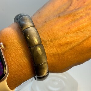 Könnte beinhalten: Ein dunkelgraues Armband mit poliertem Finish. Das Armband besteht aus rechteckigen Perlen und wird am Handgelenk getragen. Das Armband hat einen Umfang von ca. 17,8 cm.