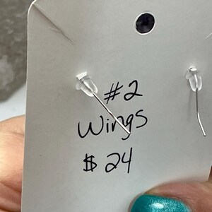 Puede incluir: Primer plano de una tarjeta blanca para exhibici&oacute;n de pendientes con el texto escrito a mano "#2 Wings 24$". La tarjeta tiene dos postes de pendientes con respaldos de pl&aacute;stico transparente. El fondo est&aacute; borroso, con una u&ntilde;a turquesa en primer plano.
