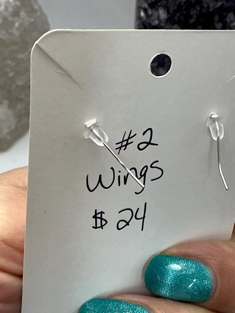Puede incluir: Primer plano de una tarjeta blanca para exhibici&oacute;n de pendientes con el texto escrito a mano "#2 Wings 24$". La tarjeta tiene dos postes de pendientes con respaldos de pl&aacute;stico transparente. El fondo est&aacute; borroso, con una u&ntilde;a turquesa en primer plano.