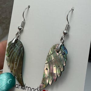 Puede incluir: Un par de pendientes hechos a mano con colgantes iridiscentes en forma de ala. Los pendientes tienen ganchos plateados y se exhiben en una tarjeta blanca con el texto "handmade with love". Las alas tienen una apariencia natural y texturizada.