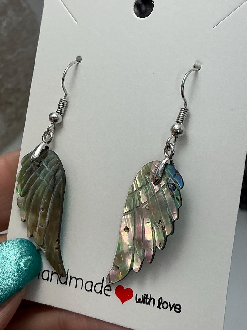 Puede incluir: Un par de pendientes hechos a mano con colgantes iridiscentes en forma de ala. Los pendientes tienen ganchos plateados y se exhiben en una tarjeta blanca con el texto "handmade with love". Las alas tienen una apariencia natural y texturizada.