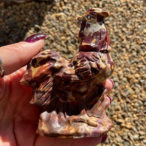 May include: A hand holds a small, intricately carved rooster figurine made of red, white, and brown stone. The rooster is standing on a base and has a detailed head and tail.