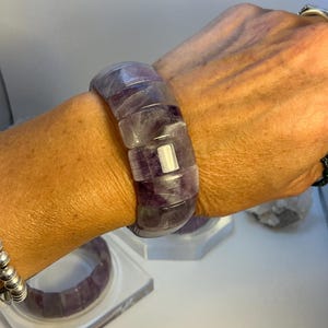 Puede incluir: Un brazalete grueso de fluorita morada y blanca. El brazalete está compuesto por piedras rectangulares pulidas. El color es una mezcla de morado translúcido y blanco. El brazalete se lleva en la muñeca. Otro brazalete es visible en el fondo.