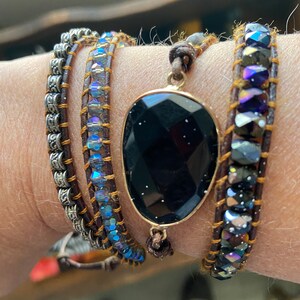 May include: A multi-strand bracelet with a large black gemstone pendant. The bracelet features brown leather cord, silver chain, and iridescent blue beads.