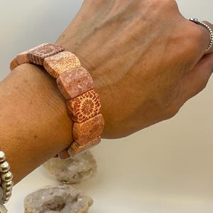 Könnte beinhalten: Ein elastisches Armband aus rechteckigen, glattkantigen Perlen mit einem markanten orange- und beigefarbenen Korallenfossil-Muster, das auf jedem einzelnen Steinsegment komplizierte, sternförmige Radialstrukturen aufweist.