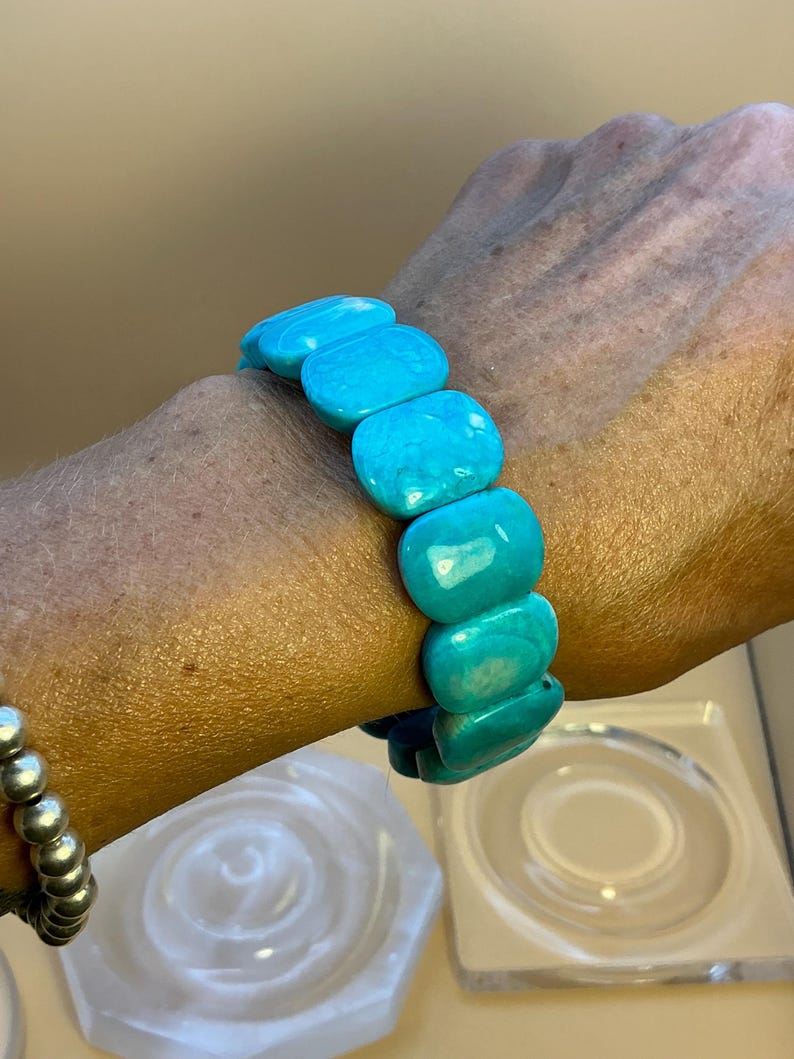 May include: A stretch bracelet composed of smooth, rectangular turquoise-colored beads with natural marbling. The beads are arranged in a single row, featuring a vibrant cyan hue with subtle white and darker blue veining throughout the polished surface.