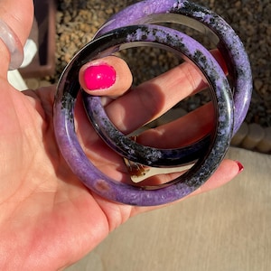 Pode incluir: Três pulseiras de pedra roxa e preta são seguradas em uma mão com esmalte rosa brilhante. As pulseiras têm uma aparência marmorizada com uma mistura de tons roxos e pretos. O fundo está desfocado, sugerindo um ambiente externo.