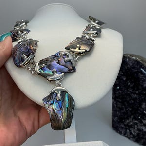 May include: Abalone shell necklace with silver-tone metal accents. The necklace features iridescent, multi-colored abalone shell pieces in shades of blue, green, and purple. The necklace is displayed on a white bust.