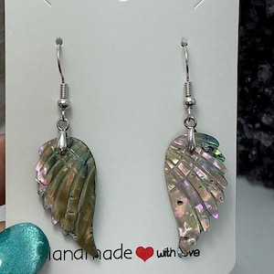 Puede incluir: Un par de pendientes iridiscentes en forma de ala con ganchos plateados. Los pendientes reflejan tonos verdes, rosas y morados. La tarjeta detr&aacute;s de los pendientes dice "handmade with love".