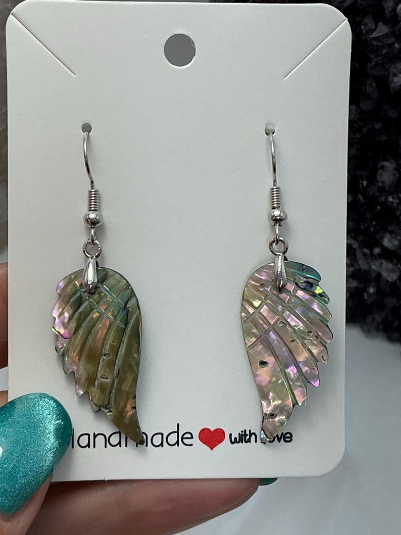 Puede incluir: Un par de pendientes iridiscentes en forma de ala con ganchos plateados. Los pendientes reflejan tonos verdes, rosas y morados. La tarjeta detr&aacute;s de los pendientes dice "handmade with love".