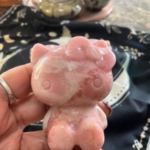 Puede incluir: Una figurita rosa de Hello Kitty tallada en cuarzo rosa. La figurita mide aproximadamente 7,5 cm de alto y tiene un acabado brillante.