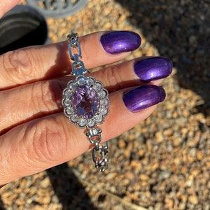 May include: A silver chain bracelet featuring a large oval amethyst gemstone surrounded by smaller clear gemstones. The bracelet is on a hand with purple nail polish.