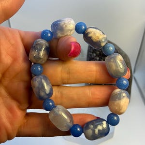 Puede incluir: Una pulsera de cuentas con piedras azules y blancas. La pulsera tiene grandes cuentas ovaladas en tonos azules y blancos, intercaladas con cuentas redondas azules más pequeñas. La pulsera está sujeta en una mano.